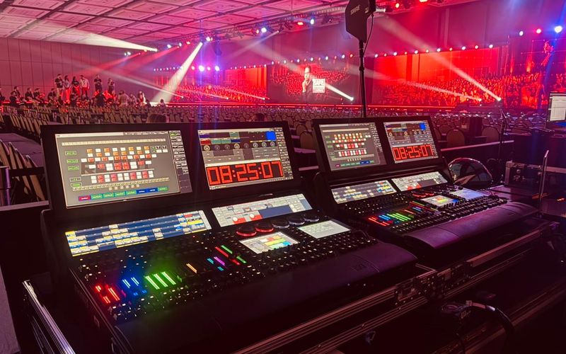 Mesa técnica de audiovisual em primeiro plano com plenária iluminada ao fundo